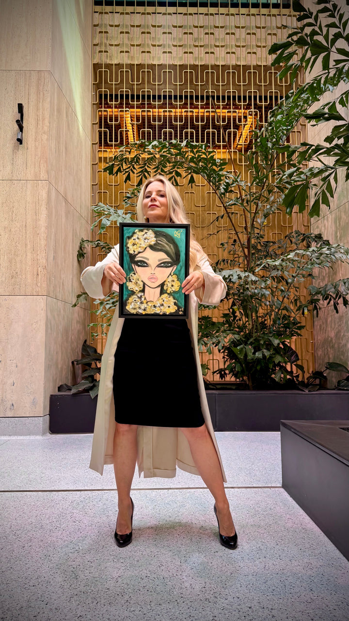 A woman stands indoors holding up a framed portrait painting of a woman with a floral crown. The background features a modern architectural interior with stone walls, a decorative gold-toned metal screen, and potted green plants.