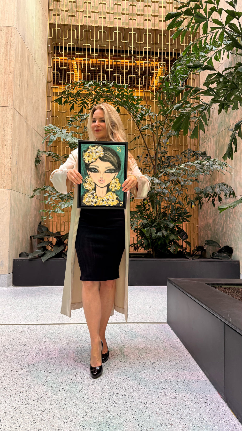 A woman stands in a modern indoor lobby holding up a framed portrait painting of a woman with dark hair and a gold floral crown. The background features a geometric gold metal screen, beige stone walls, and green potted plants.