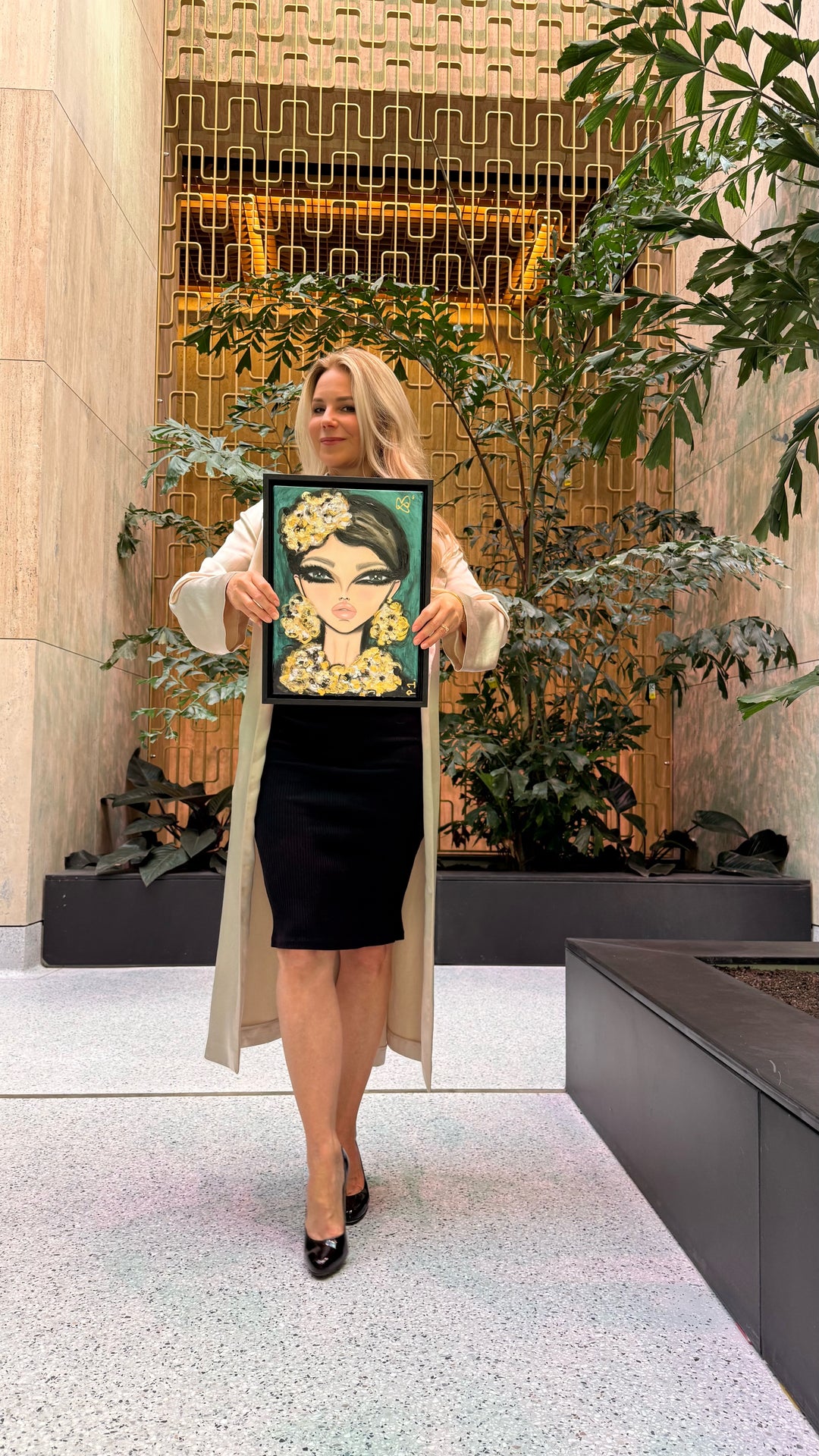 A woman stands in a modern indoor lobby holding up a framed portrait painting of a woman with dark hair and a gold floral crown. The background features a geometric gold metal screen, beige stone walls, and green potted plants.