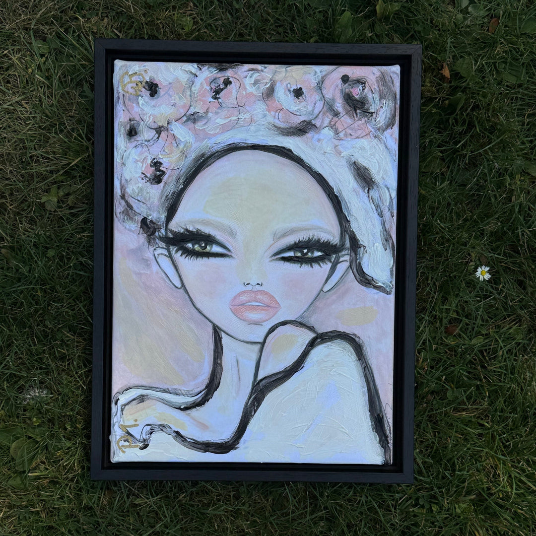 A framed portrait painting of a stylized woman with large eyes, black hair, and pink lips, placed on green grass outdoors.