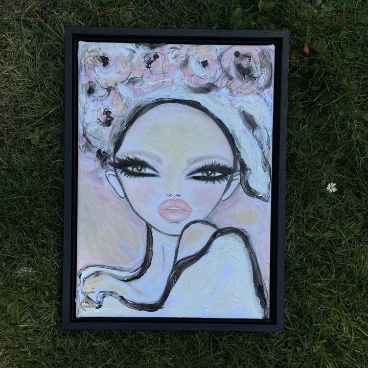 A framed portrait painting of a stylized woman with large eyes, black hair, and pink lips, placed on green grass outdoors.