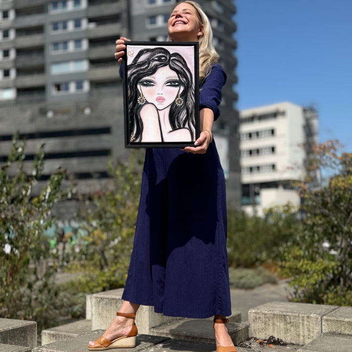 A woman in a navy blue maxi dress and tan wedge sandals stands on outdoor concrete steps holding up a framed caricature portrait painting of a woman with long black hair.