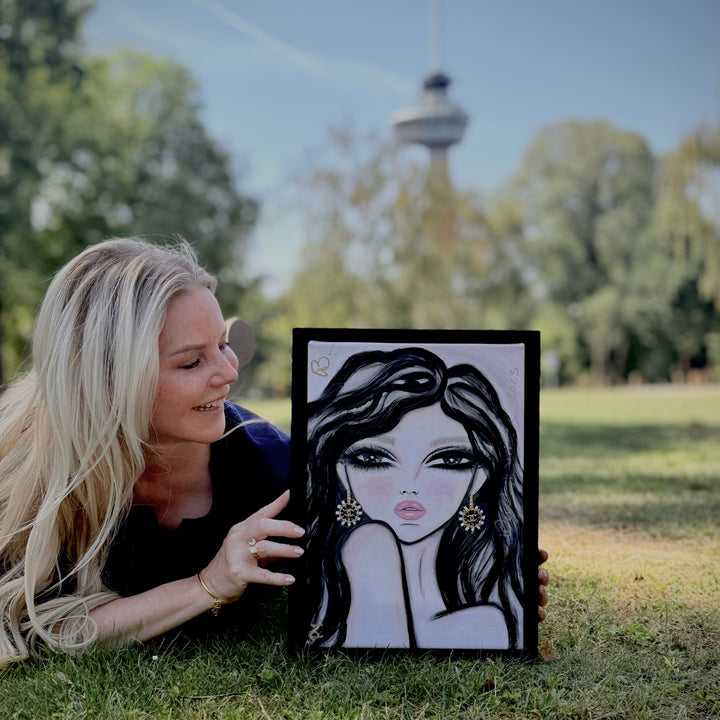A woman lying on grass in a park holds up a framed black and white portrait drawing of a woman with long dark hair and gold earrings, with the Euromast visible in the blurred background.
