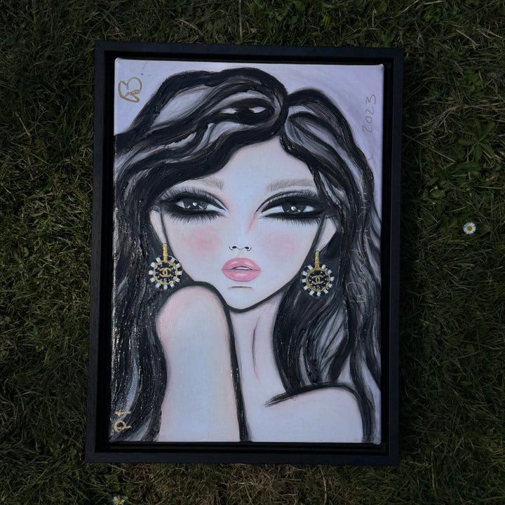 A framed portrait drawing of a woman with long dark hair, dramatic eye makeup, and gold earrings, placed on a grassy ground.