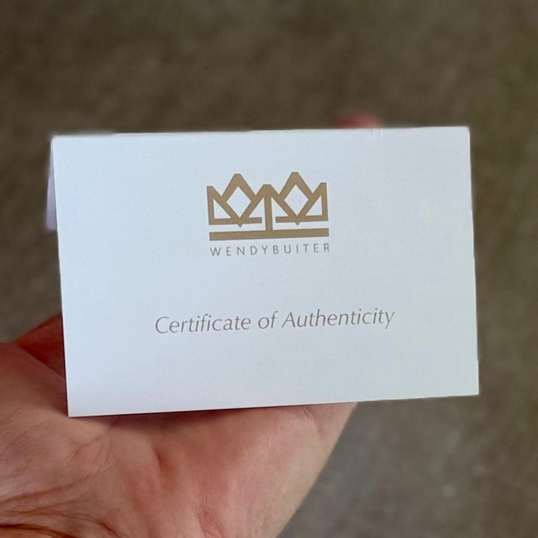 A hand holding a white card labeled 'WENDYBUITER' with a gold crown logo and the text 'Certificate of Authenticity' against a blurred brown background.