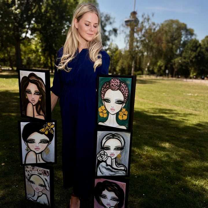 A woman in a navy blue dress stands outdoors on a grassy park lawn holding and displaying six framed stylized portrait paintings of women. The portraits feature bold black outlines, dramatic makeup, and colorful hair and background details.