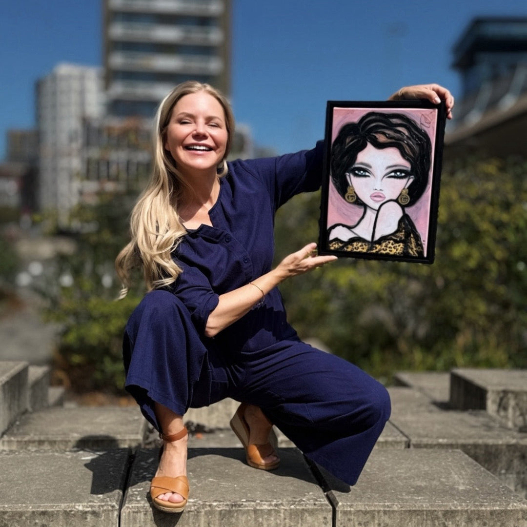 A woman in a navy blue jumpsuit crouches on concrete steps outdoors, holding up a framed portrait painting of a woman with dark hair and a pink background. Buildings and a blue sky are visible in the blurred background.