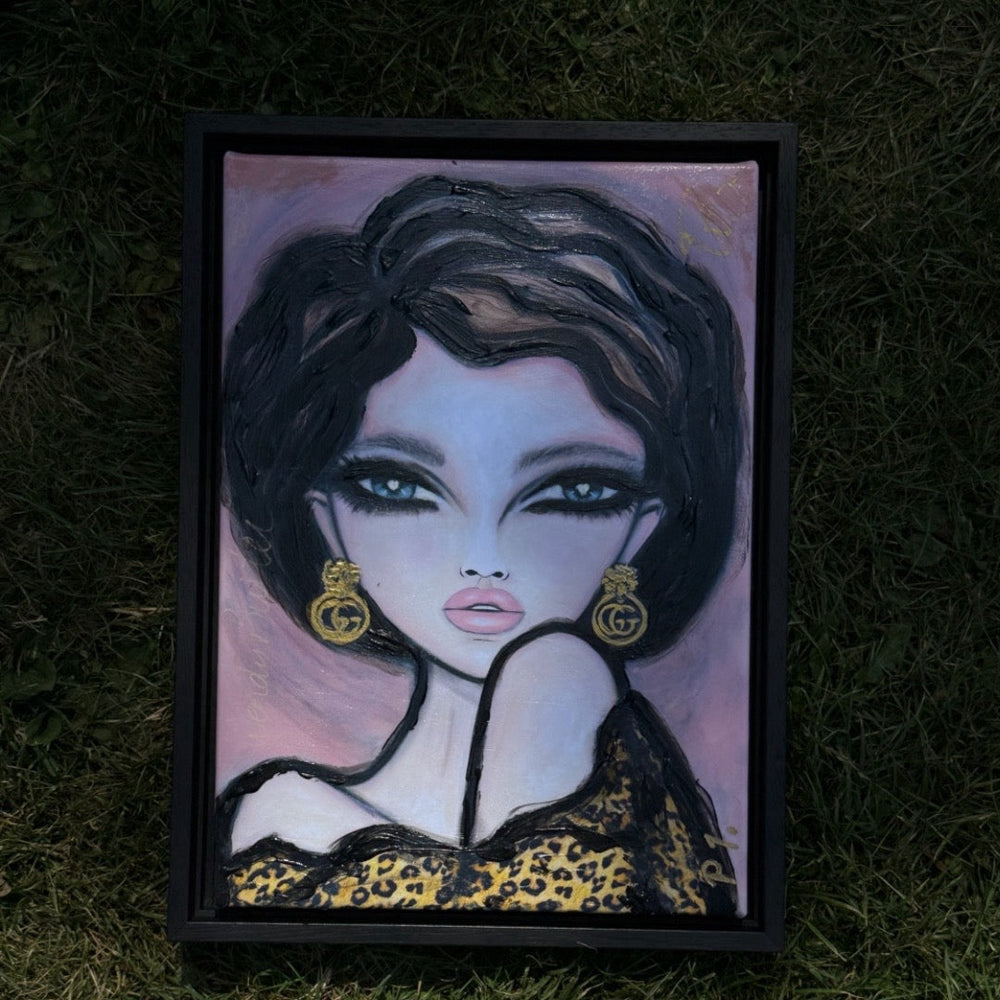 A framed portrait painting of a stylized woman with dark hair, pale skin, and gold earrings, placed on a grassy ground.
