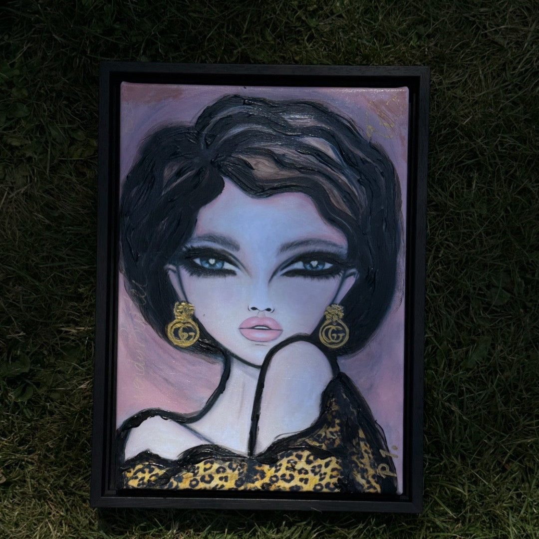 A framed portrait painting of a stylized woman with dark hair, pale skin, and gold earrings, placed on a grassy ground.