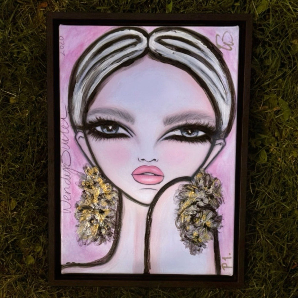 A framed portrait painting of a stylized woman with large eyes, pink lips, and textured flower-like earrings, placed on a bed of green grass.