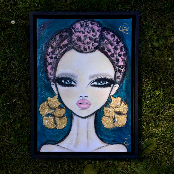 A framed portrait painting of a stylized woman with a pink leopard-print headpiece, large gold flower-shaped earrings, and a dark teal background, placed on green grass.