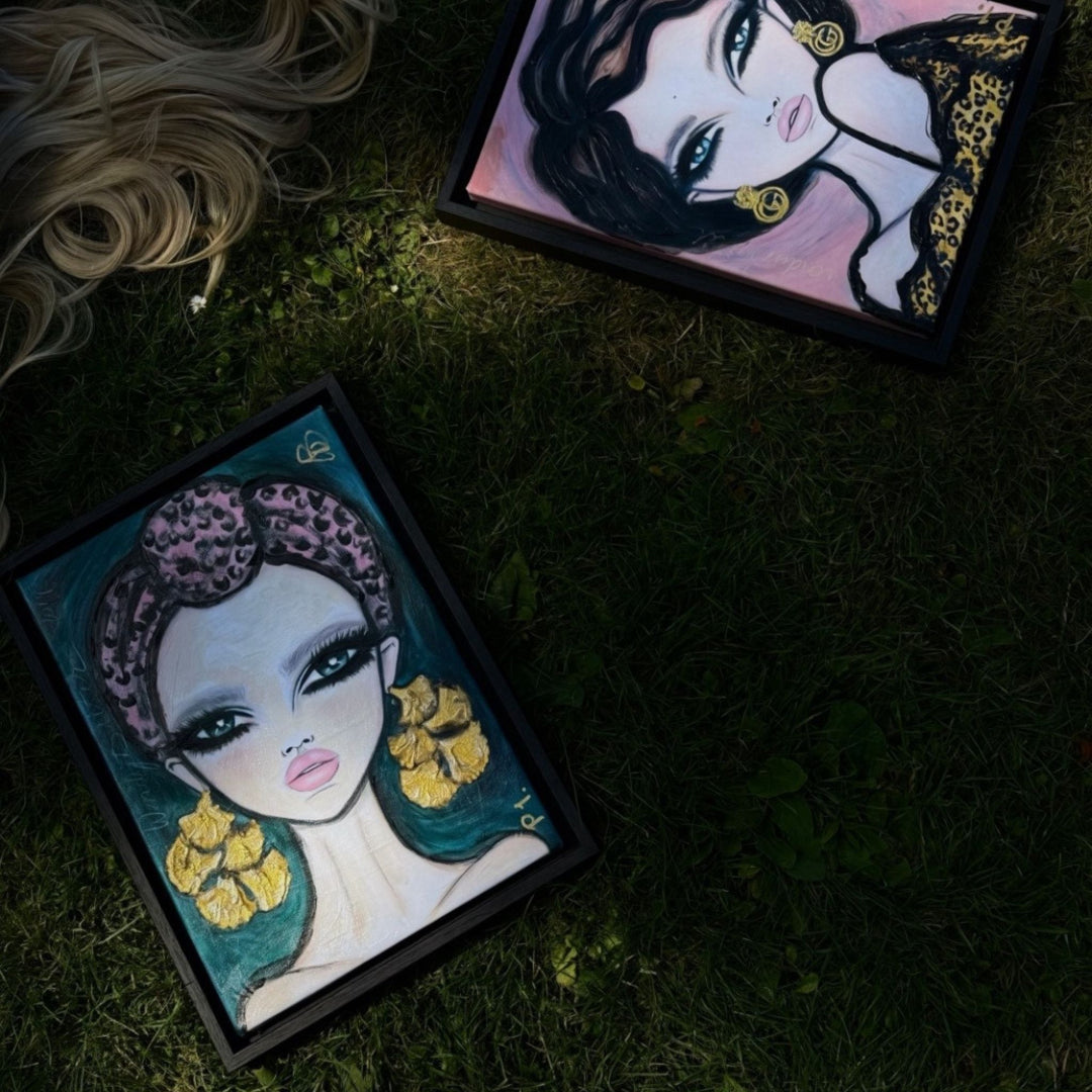 Two framed art prints depicting stylized female portraits with large eyes, leopard-print headbands, and gold earrings, placed on dark grass with a blonde woman nearby.