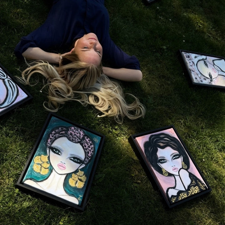 A woman lies on green grass surrounded by several framed art prints featuring stylized fashion illustrations.