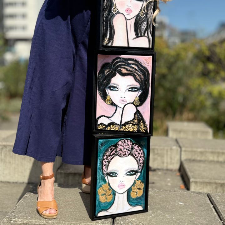 Three framed vertical portraits of stylized women with black hair and gold jewelry are stacked outdoors on a concrete surface. A person wearing a navy dress and tan sandals stands beside the frames, with blurred greenery and buildings in the background.