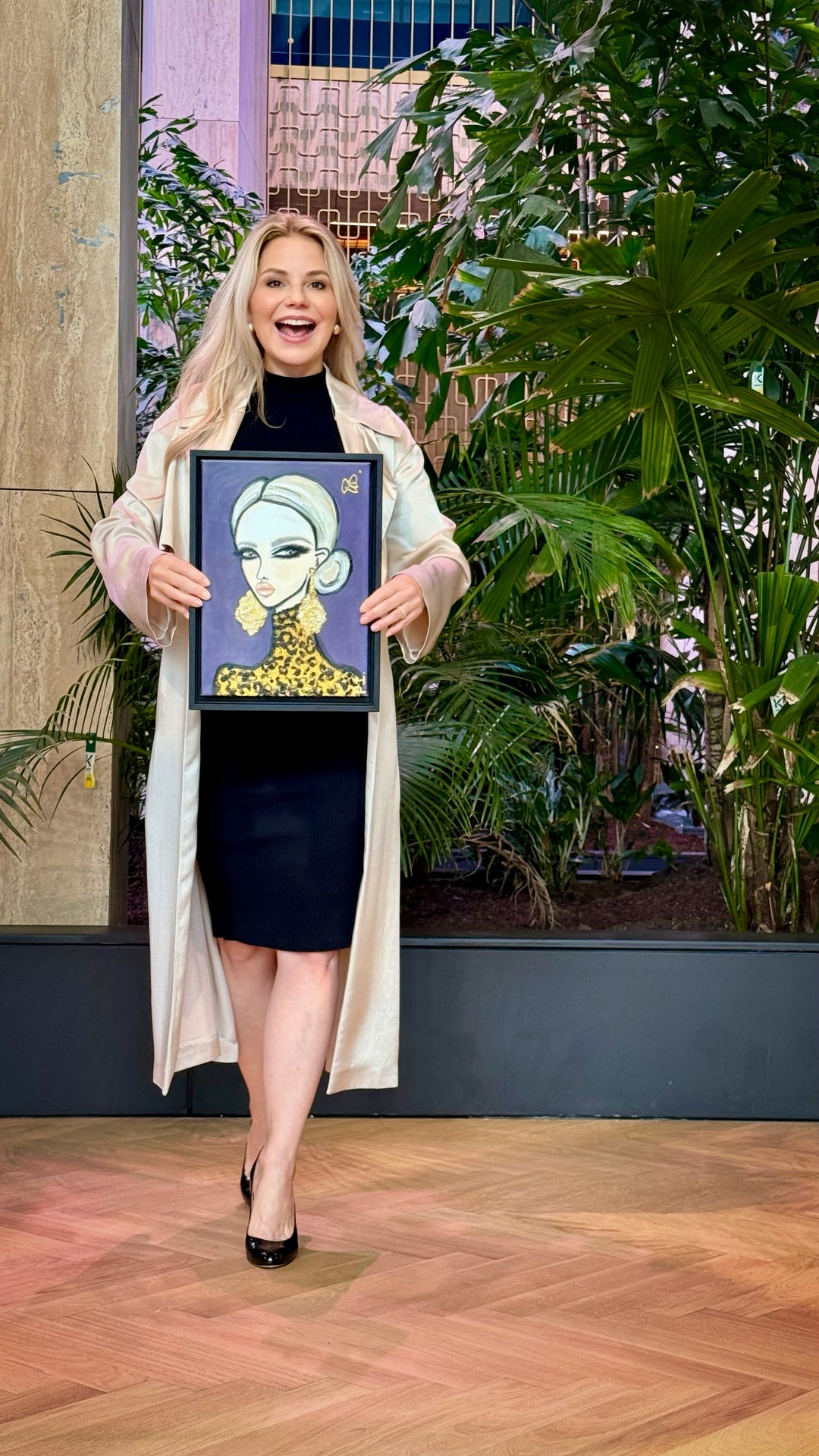 A woman stands in a studio setting with plants and wood flooring, holding up a framed art print featuring a stylized illustration of a woman with a gold collar.