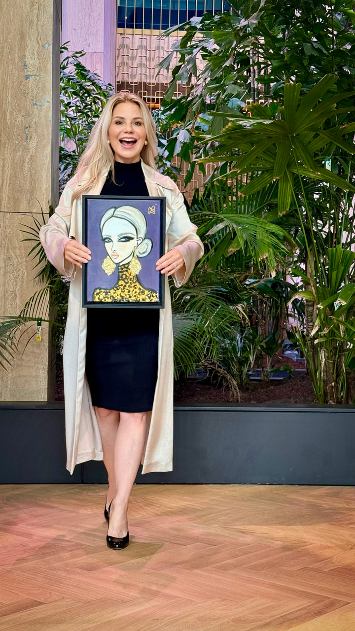 A woman stands in a studio setting with plants and wood flooring, holding up a framed art print featuring a stylized illustration of a woman with a gold collar.