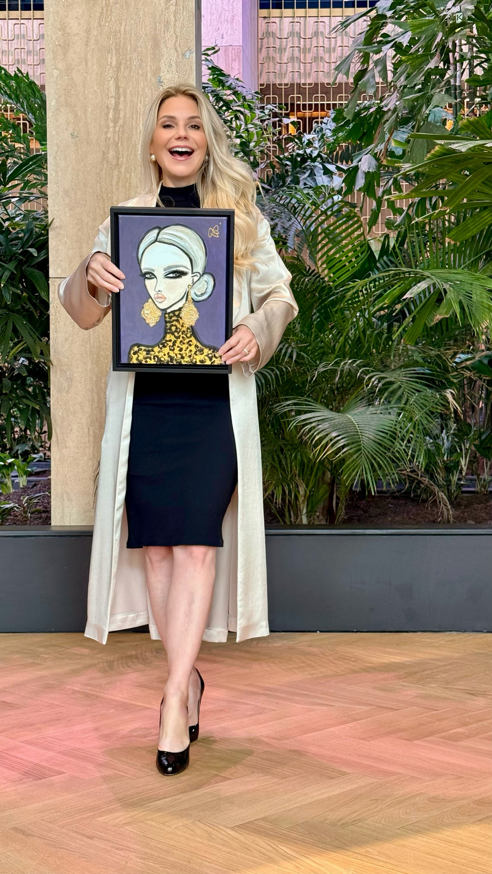 A woman stands in an indoor studio or gallery setting with plants and wood flooring, holding up a framed art print featuring a stylized portrait of a woman with gray hair, a gold patterned top, and a black background.