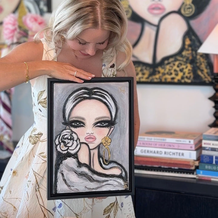 A woman in a floral dress holds up a framed portrait painting of a woman with dramatic eye makeup, a large gold hoop earring, and a rose. The background shows a bookshelf with books and another framed art piece.