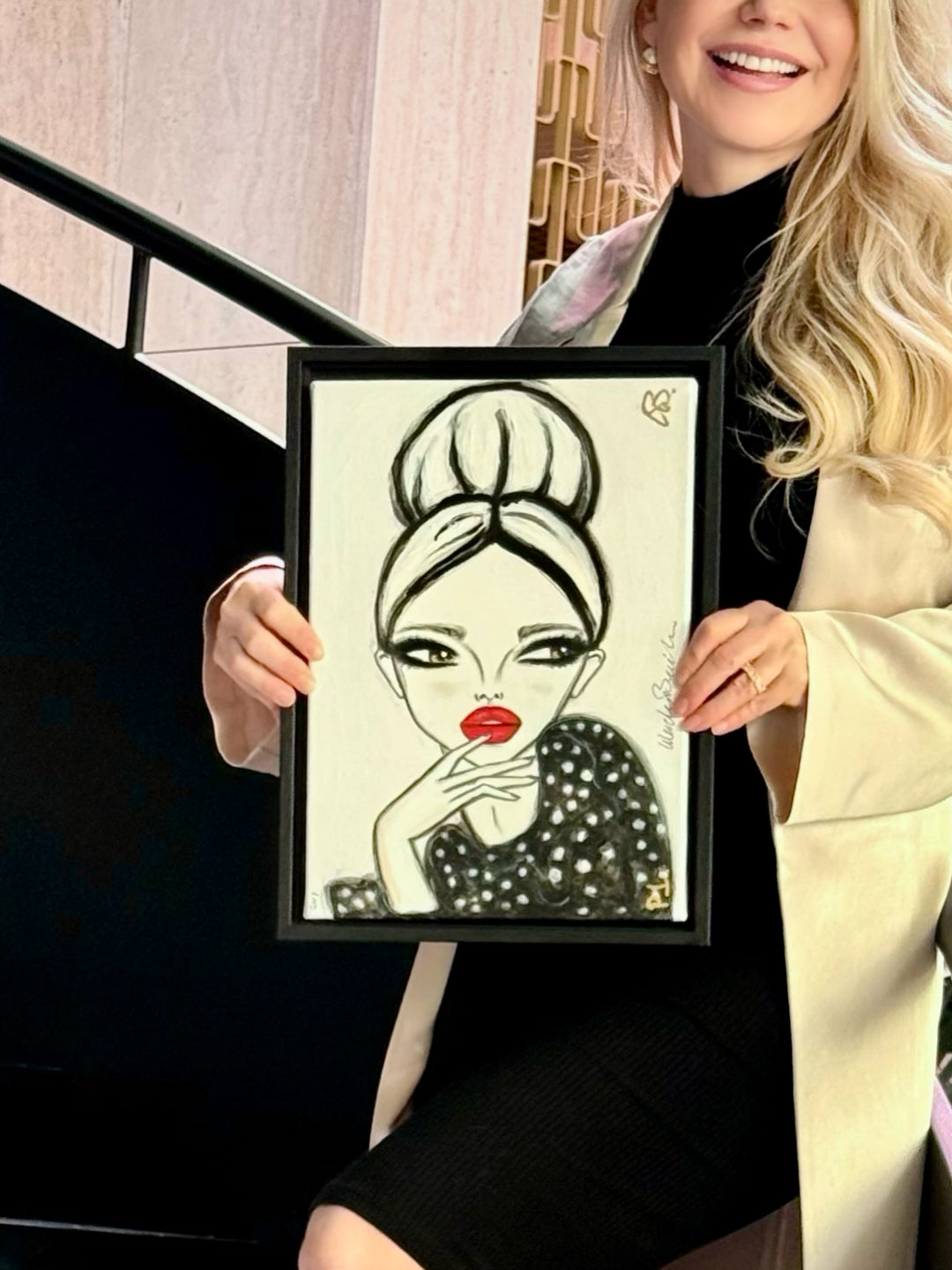 A woman holds up a framed black and white fashion illustration of a woman with red lips. The woman is wearing a black dress and a cream-colored blazer, standing in front of a staircase.