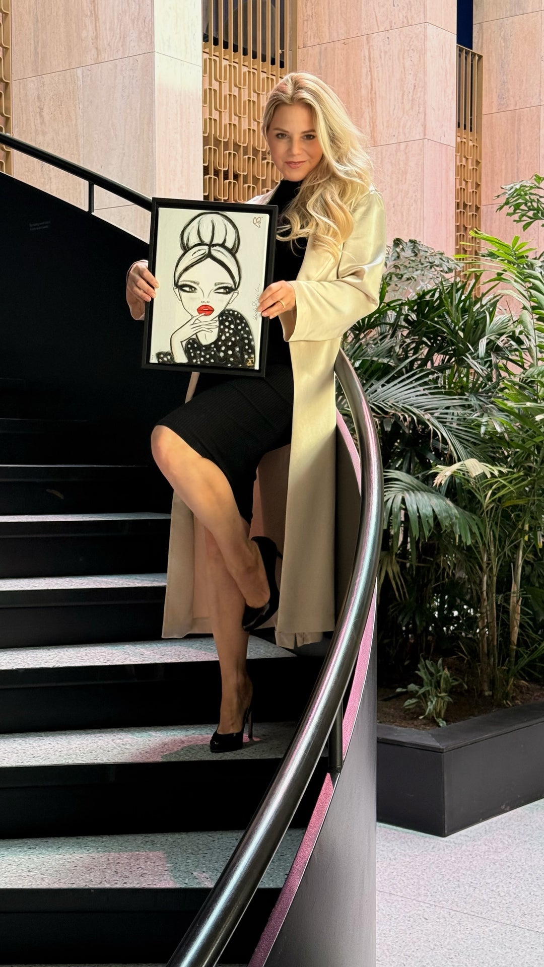 A blonde woman in a beige trench coat and black dress stands on an indoor staircase holding a framed black and white fashion illustration print with red lips.