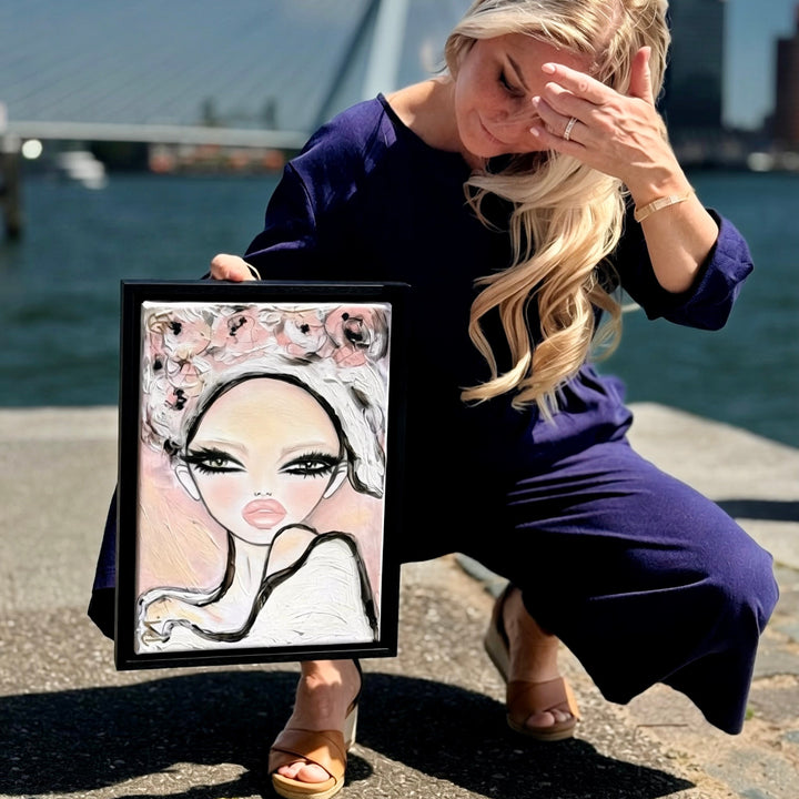 A woman in a dark purple dress kneels outdoors by a waterfront holding a framed portrait painting. The painting features a stylized woman with large eyes, black hair, and pink floral details.