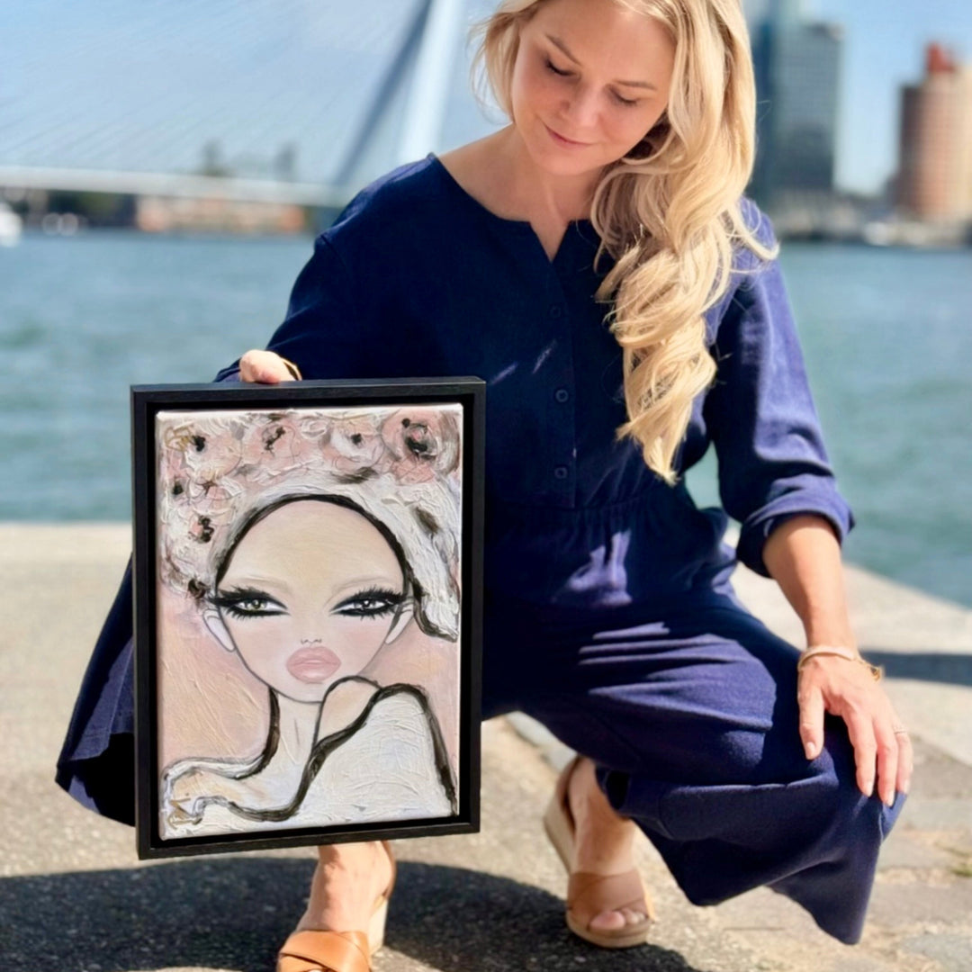 A woman in a navy blue jumpsuit crouches outdoors by the water holding up a framed portrait painting of a stylized woman with a floral headpiece.