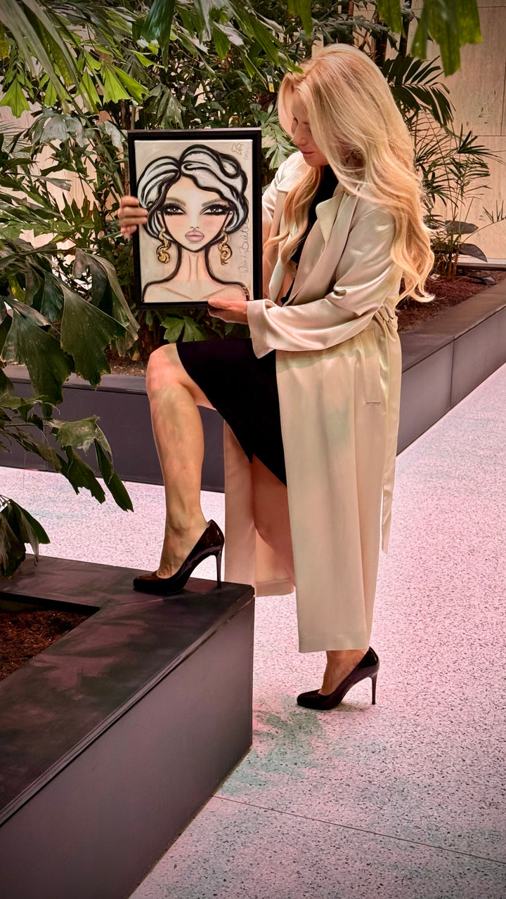 A woman in a cream satin trench coat, black dress, and black heels holds up a framed fashion illustration of a woman with wavy black hair and gold hoop earrings, posing in a modern indoor garden setting.