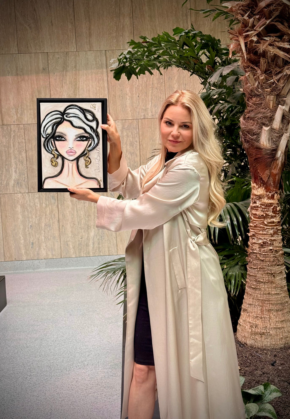 A woman in a long cream satin robe stands indoors next to a palm tree, holding up a framed fashion illustration of a woman with blonde hair and gold hoop earrings.