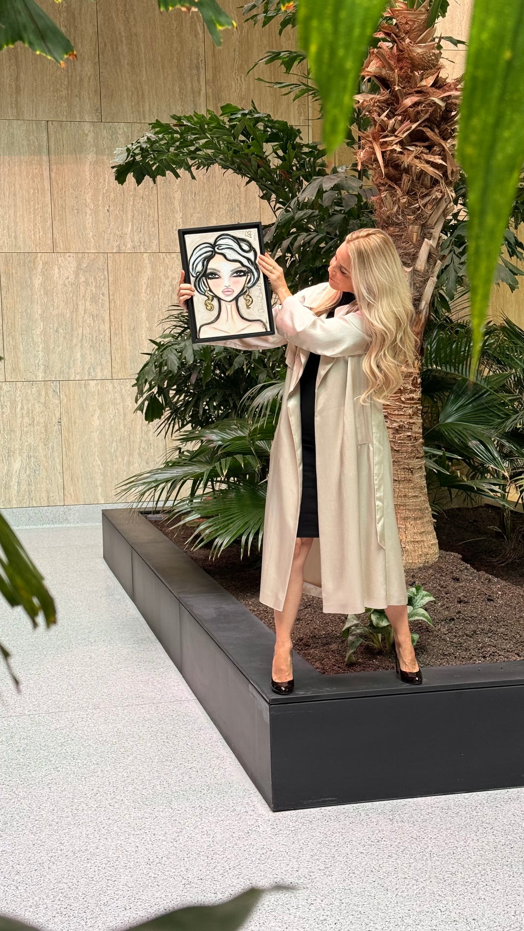 A woman in a long cream trench coat and black dress holds up a framed black and white portrait illustration in an indoor garden setting with plants and a stone wall.
