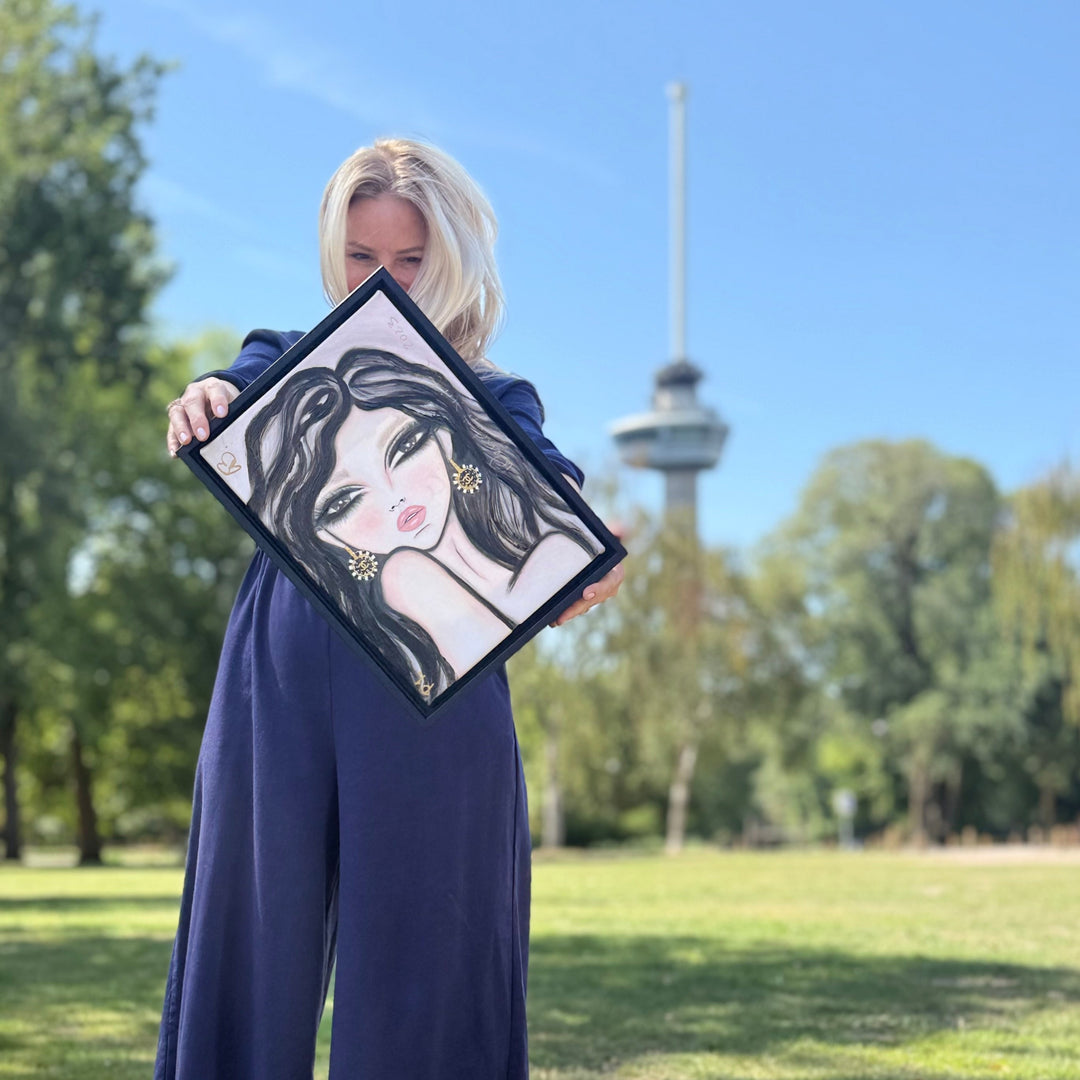 A woman in a navy blue wide-leg jumpsuit stands on a grassy lawn holding up a framed art print of a stylized woman with long black hair. The background shows a park with trees and a tower under a clear blue sky.
