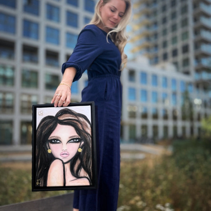 A woman in a navy blue jumpsuit stands on a bench in an outdoor urban setting, holding up a framed portrait illustration of a woman with dark hair and gold earrings.