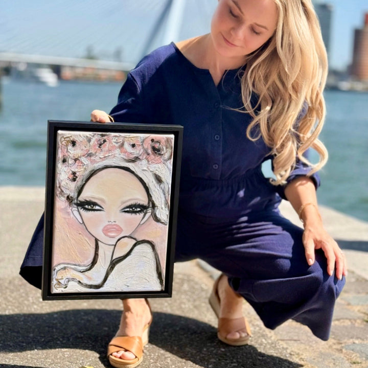 A woman in a navy blue jumpsuit kneels outdoors by the water holding a framed custom portrait painting. The painting features a stylized woman with a floral headpiece.