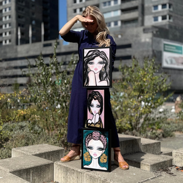 A woman in a navy blue dress stands on outdoor concrete steps holding a vertical stack of three framed art prints featuring stylized portraits of women. The background shows tall urban buildings and greenery.