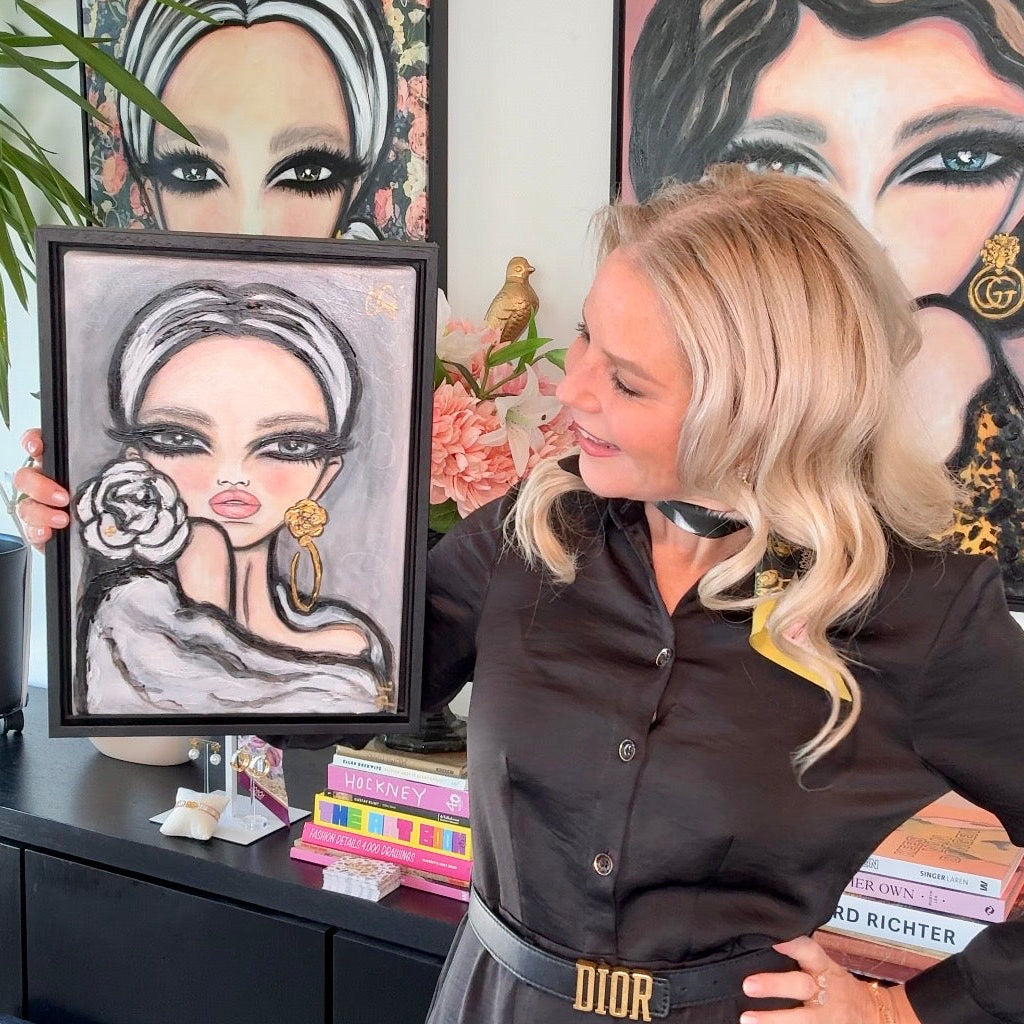 A blonde woman in a black button-up shirt and gray skirt with a gold DIOR belt holds up a framed portrait painting of a woman with a rose. She is standing in a room with other framed portraits on the wall behind her.