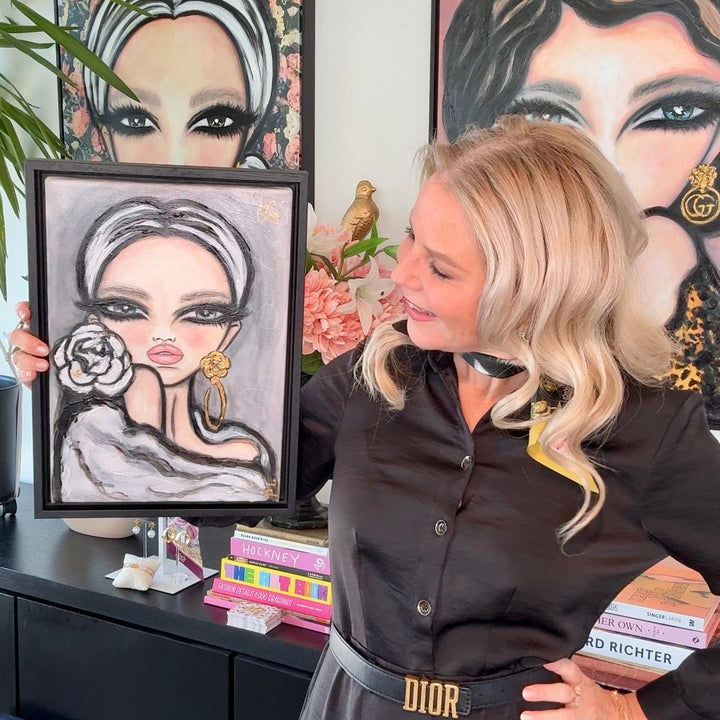 A blonde woman in a black button-up shirt and gray skirt with a gold DIOR belt holds up a framed portrait painting of a woman with a rose. She is standing in a room with other framed portraits on the wall behind her.