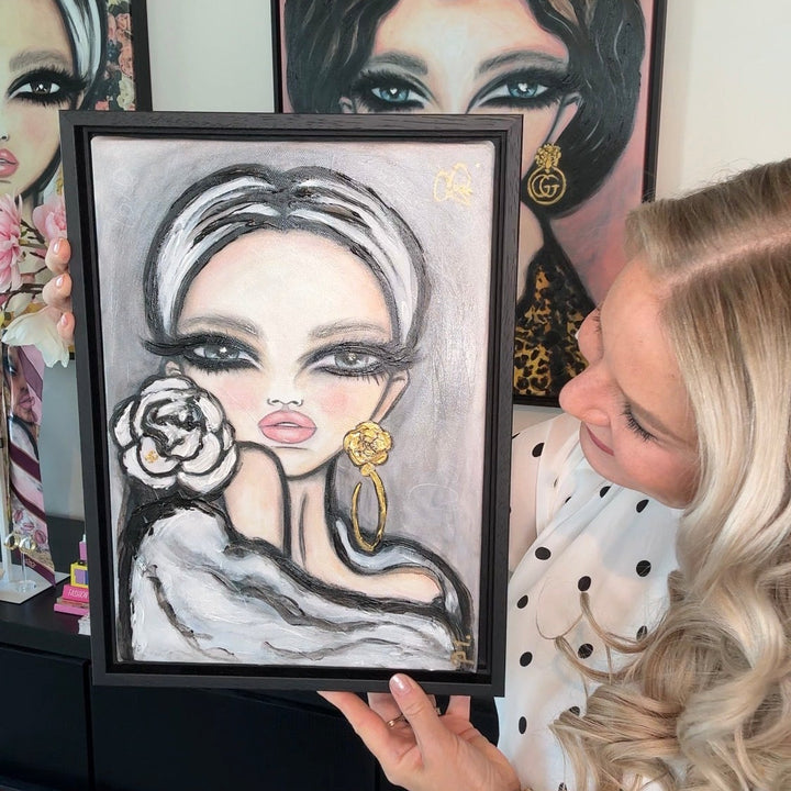 A woman in a white blouse with black polka dots holds up a framed portrait painting of a woman with gray hair and gold earrings. Other framed portraits are hanging on the wall behind her.