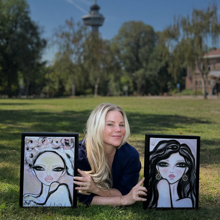 A woman lies on a grassy lawn outdoors, holding up two framed caricature portraits of herself. The background shows a park with trees and a tall tower under a blue sky.