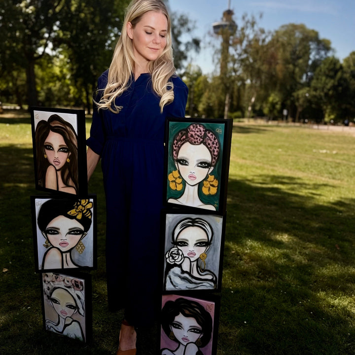 A woman in a dark blue dress stands outdoors on a grassy park lawn holding three vertical panels displaying stylized fashion illustrations of women's faces.