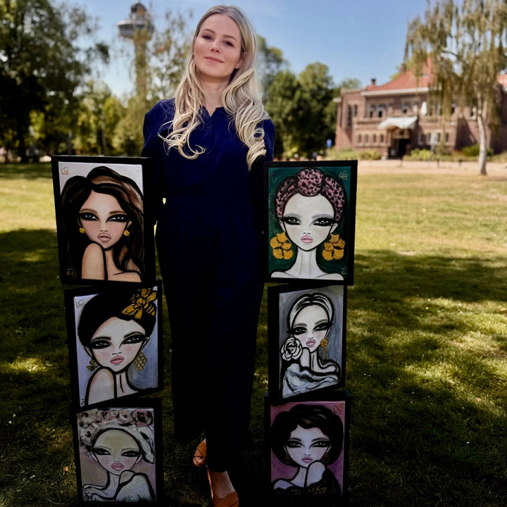 A woman stands outdoors on a grassy lawn holding two tall stacks of square paintings featuring stylized illustrated female portraits. The background shows trees, a building, and a blue sky.